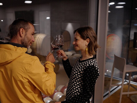 Un uomo e una donna  stanno alzando i loro calici di vino rosso, sorridendosi a vicenda. Si trovano all'interno di una cantina moderna con pareti di vetro e pavimenti in legno. Dietro l'uomo, sono visibili delle botti di vino accatastate in un'altra stanza. L'atmosfera è intima e l'illuminazione è soffusa.