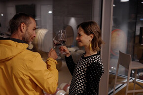 Un uomo e una donna  stanno alzando i loro calici di vino rosso, sorridendosi a vicenda. Si trovano all'interno di una cantina moderna con pareti di vetro e pavimenti in legno. Dietro l'uomo, sono visibili delle botti di vino accatastate in un'altra stanza. L'atmosfera è intima e l'illuminazione è soffusa.