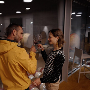 Un uomo e una donna  stanno alzando i loro calici di vino rosso, sorridendosi a vicenda. Si trovano all'interno di una cantina moderna con pareti di vetro e pavimenti in legno. Dietro l'uomo, sono visibili delle botti di vino accatastate in un'altra stanza. L'atmosfera è intima e l'illuminazione è soffusa.