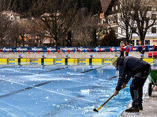 Staff members work at the edge of an outdoor pool, using shovels to break and remove a thin layer of ice from the water's surface. This image shows the pool being prepared for the Ice Swimming Championships.