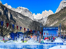 Athletes compete in the backstroke in an outdoor pool during the Ice Swimming World Championships. The low-angle shot, taken at water level, shows the pool surrounded by ice and the imposing Dolomite mountains providing a backdrop for the competition.