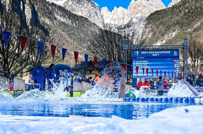 Atleti gareggiano a dorso in una piscina all'aperto durante i Mondiali di "Ice Swimming". La ripresa, a pelo d'acqua, mostra la piscina circondata da ghiaccio e le imponenti montagne dolomitiche che fanno da sfondo alla competizione.