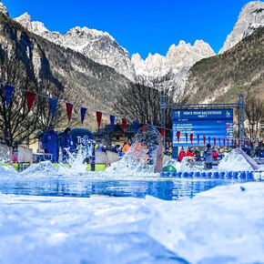 Atleti gareggiano a dorso in una piscina all'aperto durante i Mondiali di "Ice Swimming". La ripresa, a pelo d'acqua, mostra la piscina circondata da ghiaccio e le imponenti montagne dolomitiche che fanno da sfondo alla competizione.
