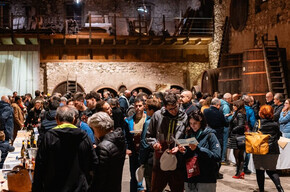 Inside a historic winery with a very high ceiling and exposed wooden beams, a wine tasting event is taking place. On the sides of the room, there are large wooden barrels, and nearby are tasting stations managed by local producers and other winemakers invited to the event. A large group of people is enjoying the experience, chatting with the producers and sipping wine.
