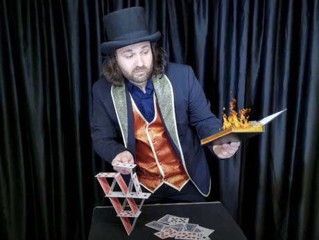 Nell'immagine si vede il mago mentre sfoglia un libro infuocato e costruisce un castello di carte