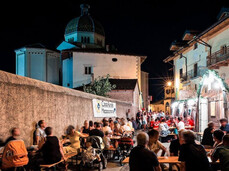 Many people are sitting at long country-style tables along a historic alley in Mezzocorona. It’s evening, everyone is having dinner, and in the background you can glimpse the inner courtyard of a house, where a local association is probably cooking for all the guests.