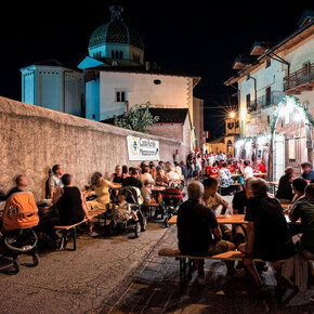 Many people are sitting at long country-style tables along a historic alley in Mezzocorona. It’s evening, everyone is having dinner, and in the background you can glimpse the inner courtyard of a house, where a local association is probably cooking for all the guests.