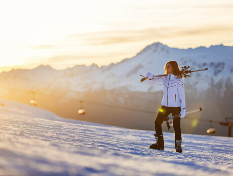 Woman carrying skis on shoulder in snowy mountain at sunset