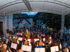 Una fotografia mostra un'esibizione di un'orchestra di fisarmoniche su un palco all'aperto, in una piazza di paese. I musicisti, quasi tutti uomini e vestiti con abiti tradizionali marroni e camicie chiare, sono seduti con i loro strumenti e spartiti. Il palco è coperto da una struttura metallica e sullo sfondo si vede una folla di spettatori seduti e in piedi. L'ambiente circostante include edifici antichi, una chiesa sulla sinistra e, più lontano, tende illuminate e montagne scure sotto un cielo crepuscolare. L'atmosfera è festosa e vivace.