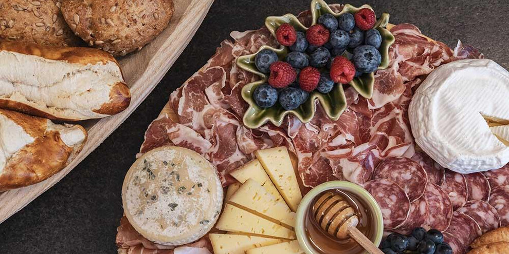 Un vassoio di salumi e formaggi con frutta fresca e miele. Accanto si trovano deliziosi pani assortiti.